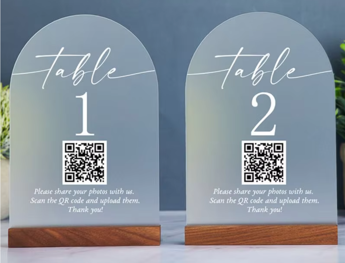 Two arched frosted glass table number signs on wooden bases, labeled table 1 and table 2 with QR codes and photo-sharing text, shown against a soft blue-gray background.
