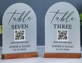 Two arched acrylic table numbers for wedding guests, one labeled 'Table Seven' and the other 'Table Three', each with a QR code and the names 'Sophie & David' on a wooden base, displayed on a marble surface against a dark gray background.