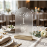 A frosted acrylic table number sign reading 'table 1' with a QR code and personalized text, displayed on a light wood base on a decorated wedding table.