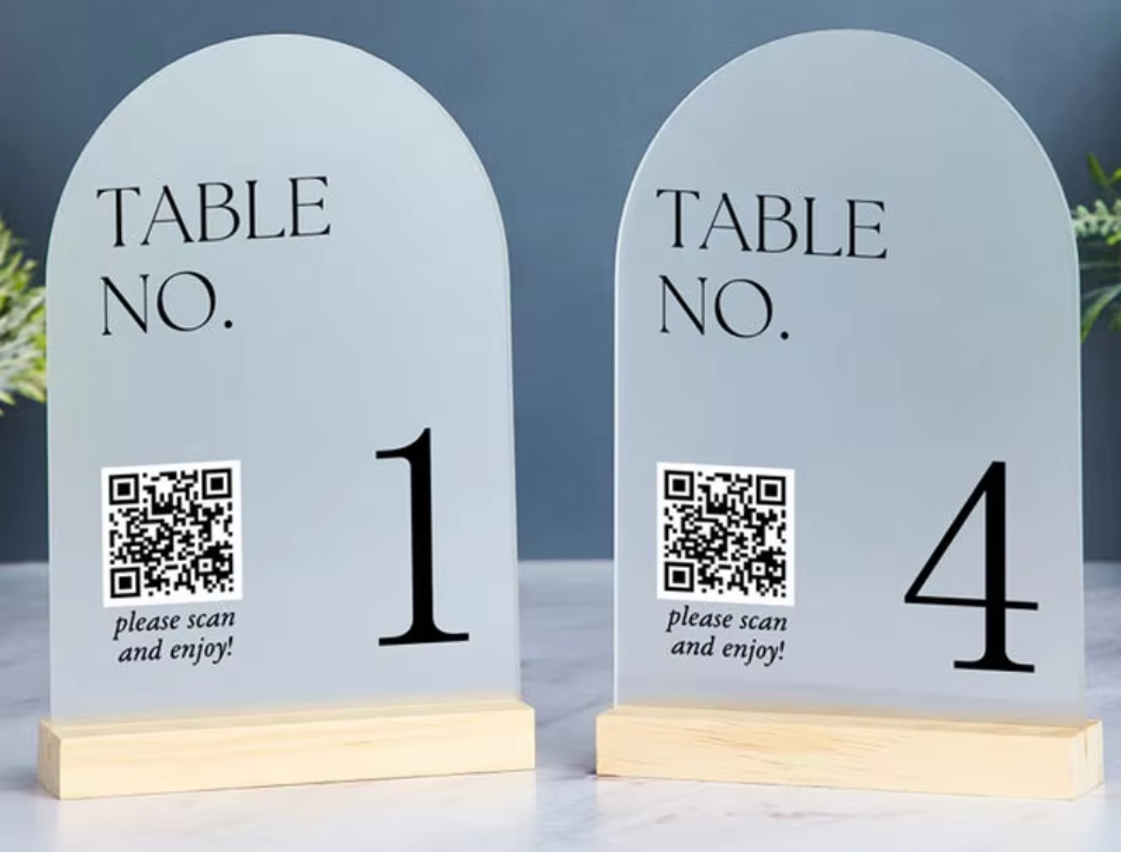 Two arched table number signs on light wood bases, one labeled 1 and the other 4, with QR codes and the text 'TABLE NO. please scan and enjoy!' on a tabletop against a blue-gray background.