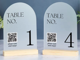 Two arched table number signs on light wood bases, one labeled 1 and the other 4, with QR codes and the text 'TABLE NO. please scan and enjoy!' on a tabletop against a blue-gray background.