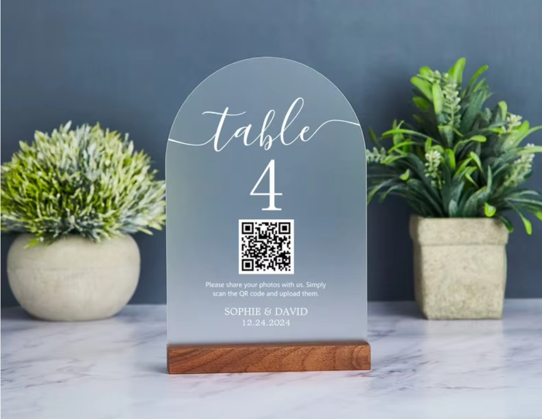 A frosted acrylic table number sign for 'table 4' with a QR code and personalized text, displayed on a wooden base on a marble surface with potted plants in the background.