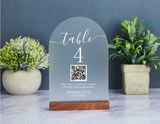 A frosted acrylic table number sign for 'table 4' with a QR code and personalized text, displayed on a wooden base on a marble surface with potted plants in the background.