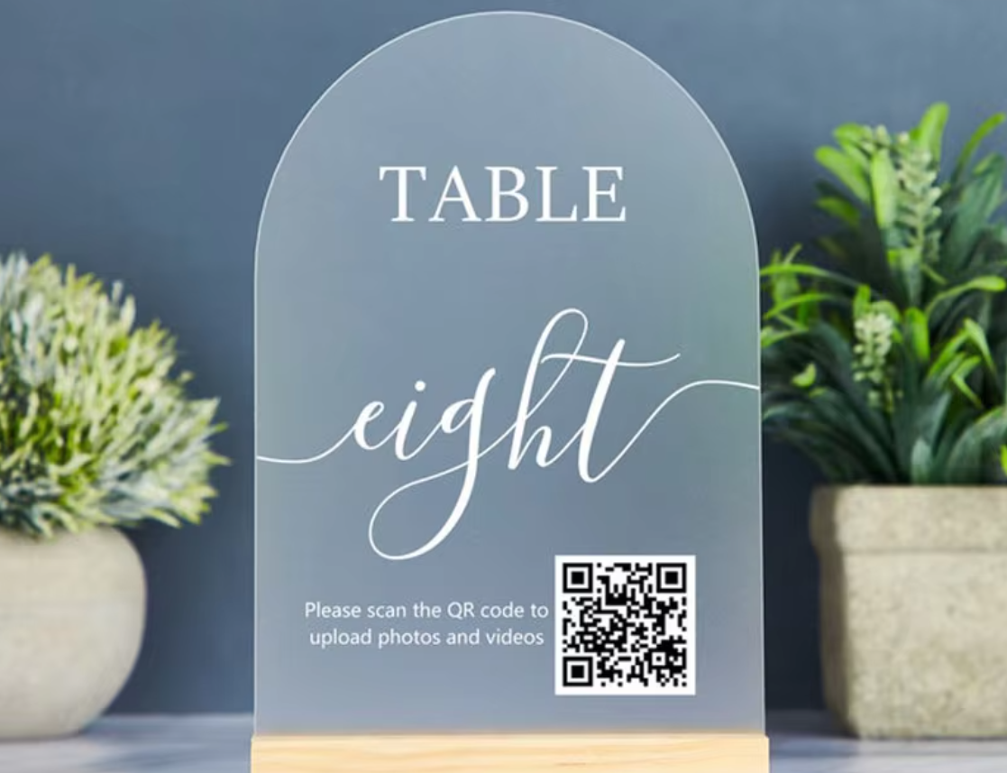 Arched acrylic table number sign reading 'TABLE eight' with a small QR code and text to upload photos, displayed on a wooden base between two potted plants against a dark blue wall.